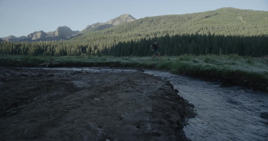 A person runs alongside a creek with a forest and mountains in the background