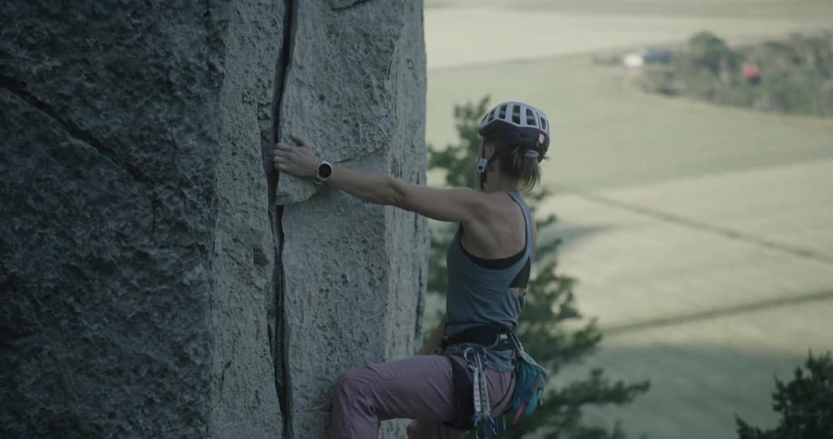 Climber wearing a helmet and harness climbs a vertical rock wall using a crevice as support with an open field in the background