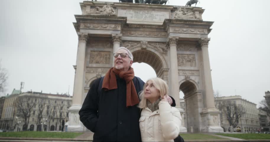 An elderly woman and man walk arm in arm near a landmark arch