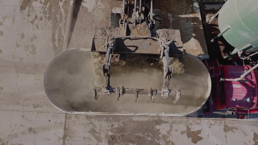 A tractor adds a large scoop of hay into a cement mixer containing water and grain on a farm