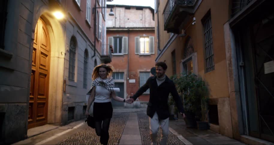 A man and a woman holding hands are running happily in an alley between large old houses
