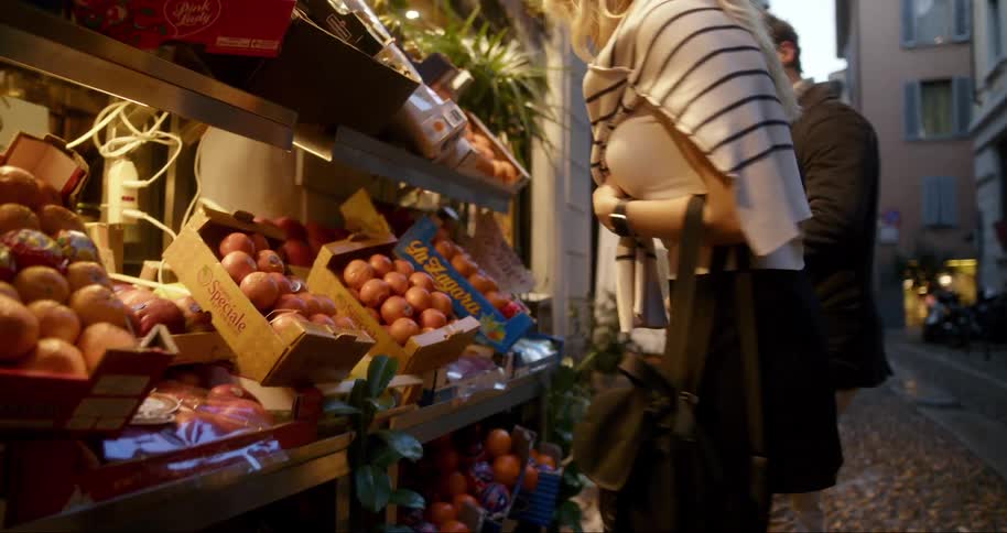 The backs of a man and a woman who are walking down an alley and then stopping at a small market with some fruit on the shelf and the woman gives the man an onion to smell and he is happy