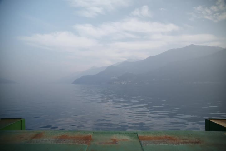 A calm lake with distant mountains is seen from a ferry deck on a hazy day