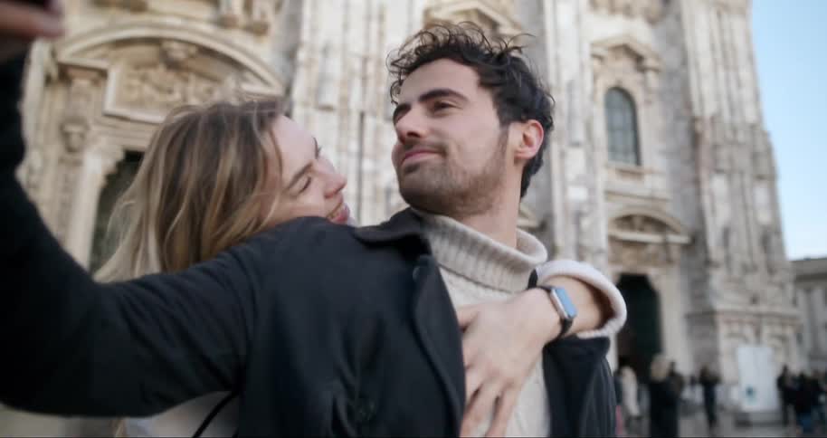 A woman smiles as she hugs a man who is taking a picture with a cellphone