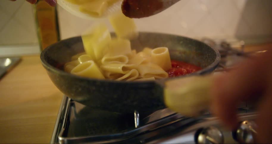 Someone pours cooked pasta into a pan of red sauce on a stovetop while another person stirs the sauce with a wooden spoon