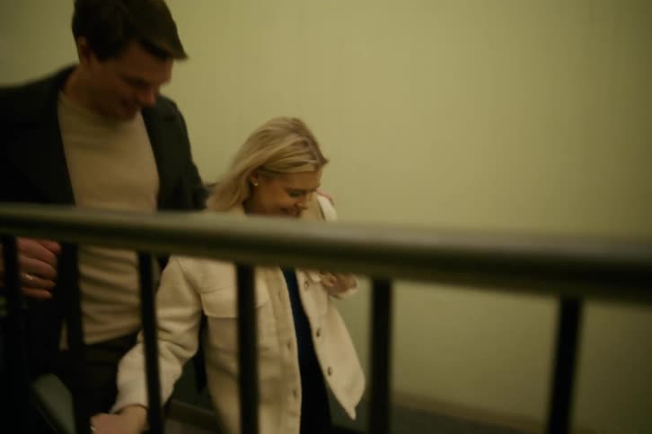 A couple walking down a staircase with a metal railing one wearing a lightcolored jacket and the other in a dark jacket and light shirt