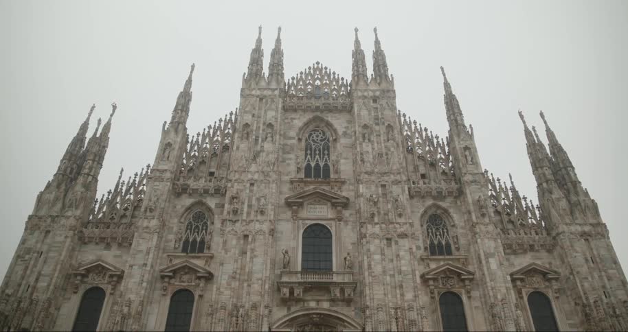 A lowangle shot shows the ornate facade of the milan duomo cathedral with multiple spires against a gray overcast sky