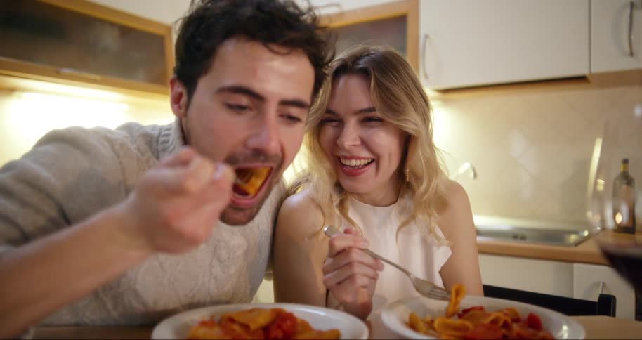 A young happy couple sits at a kitchen table eating pasta from white plates with glasses of red wine in front of them