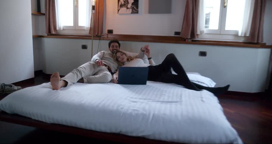 A young couple lies in bed in a neatly ordered bedroom looking at a laptop and cuddling comfortably