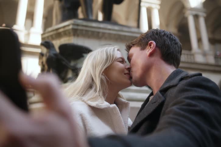 A young happy couple kiss while taking a selfie in front of an ornate building