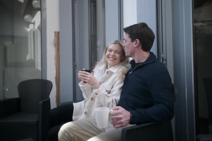 A young happy couple wearing coats sit outside a house with cups of hot beverage as the man kisses the woman on the forehead both smiling happily