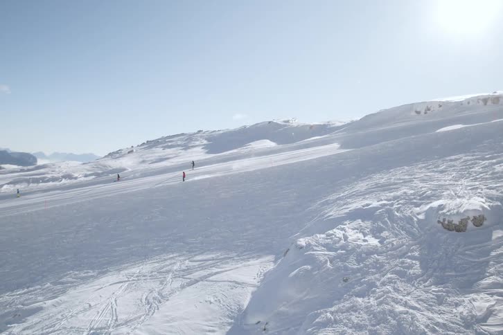 A wide shot of a snowy mountain slope with a few small figures descending on skis under a bright clear sky