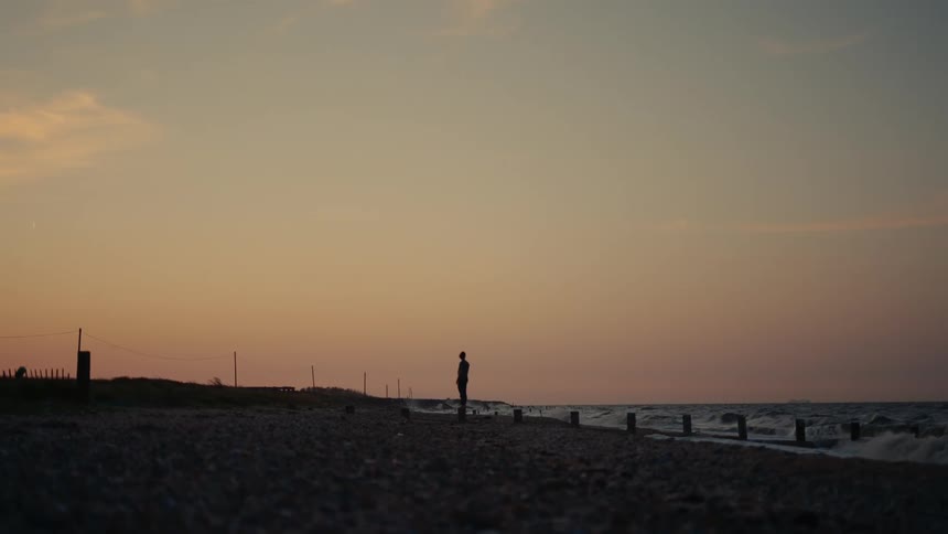 A silhouetted figure stands on a pebbled beach at dusk jumping over wooden poles buried in the sand