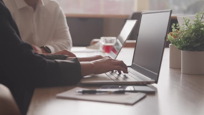 Two people are working on laptops at a desk with a plant a notepad and a pen