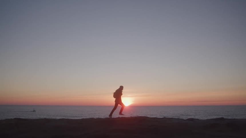 Backpacker silhouetted against a sun sitting on the red tinted horizon runs on shore