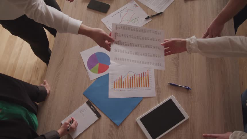 A diverse group of people are collaborating on business matters around a table scattered with charts graphs a tablet and notes