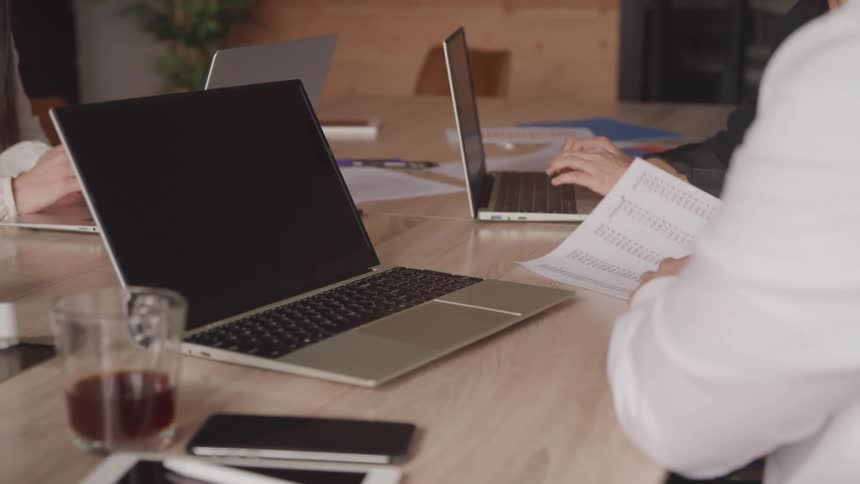 Three people sitting at an office desk with laptops open and sharing documents