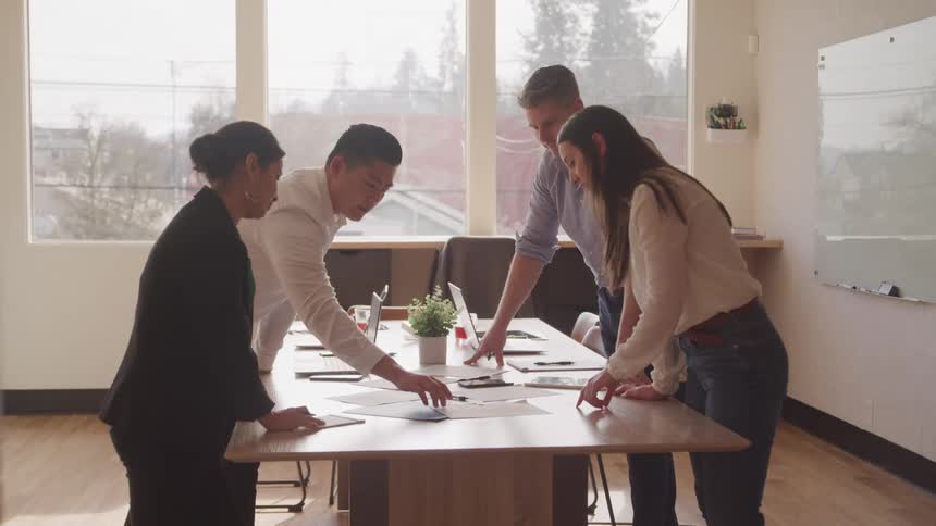 Four professionals gathered around a wooden table in a modern office engaged in a collaborative discussion as they review documents and use laptops with natural light streaming through large windows