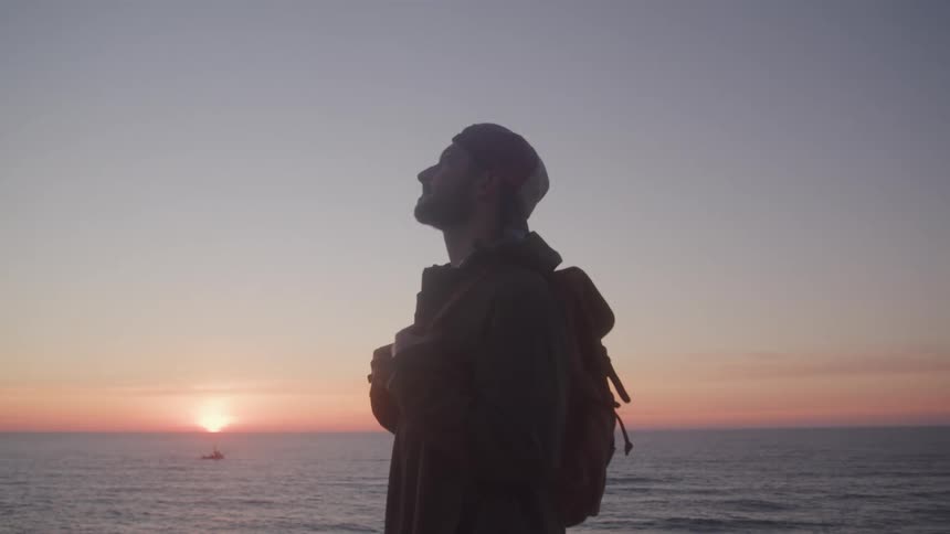 Man in cap and backpack looking up while standing in outdoor seaside setting