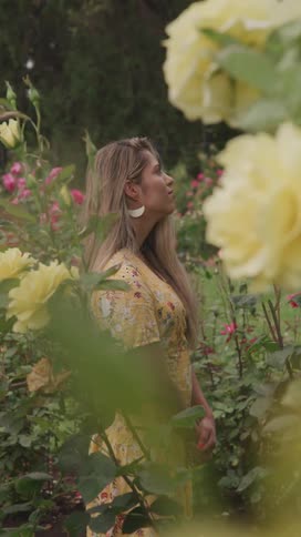 A blonde woman in a yellow dress is standing in a field of yellow flowers and then looks ahead with a subtle smile