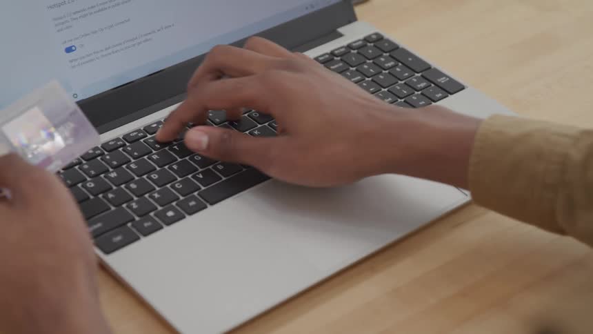 A person is using a laptop typing on the keyboard with one hand while holding a credit card in the other hand
