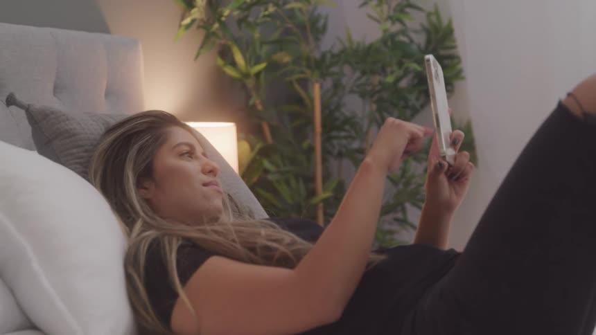A woman is laying on a bed in a bedroom and touching the screen of her cellphone