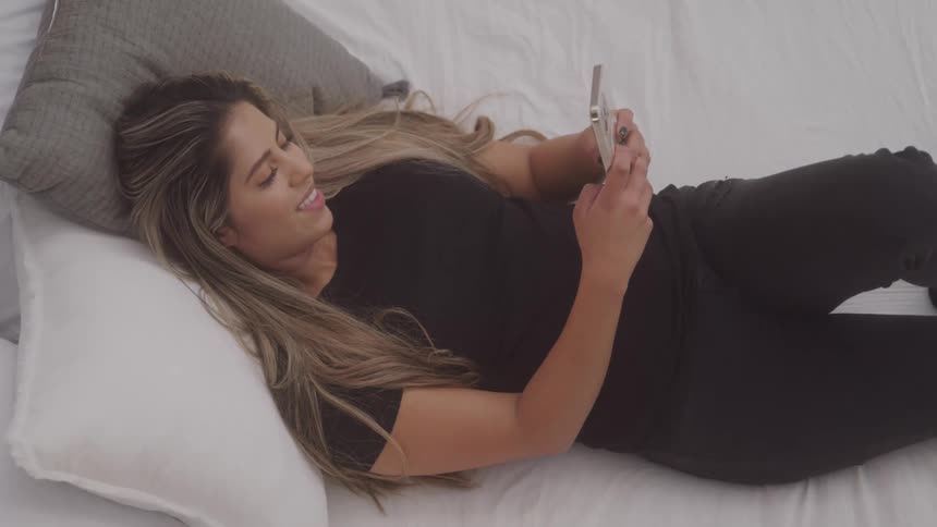 A young woman lies on a bed while typing on her smartphone