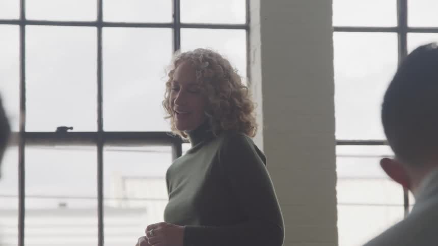 A woman stands next to a whiteboard in a welllit room gesturing with a marker in hand as she explains a concept or presents information to a group seated nearby