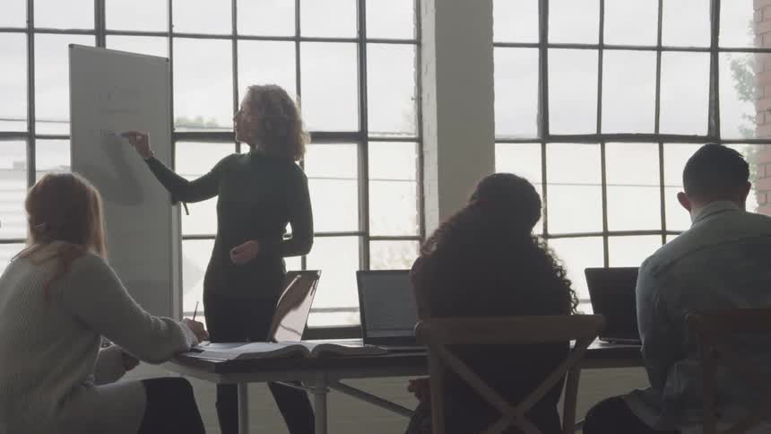 Woman standing writing on whiteboard for three other people who are sitting at a table with laptop