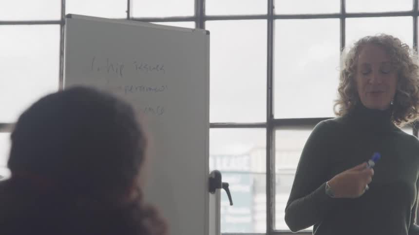 A woman stands next to a whiteboard in a welllit room gesturing with a marker in hand as she explains a concept or presents information to a group seated nearby