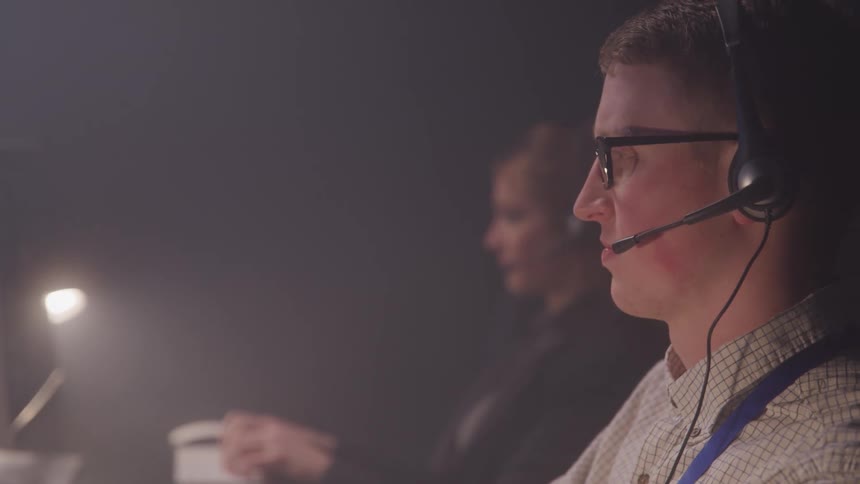 Coworkers work at a desk in a dimly lit room focused on their computers suggesting a call center or tech support setting