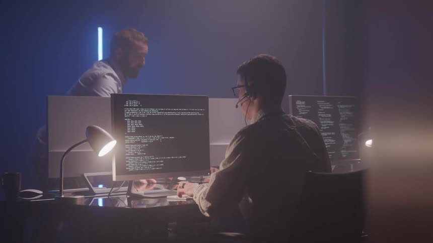People working at desks in a dimly lit room focused on computers resembling a call center or tech support setting