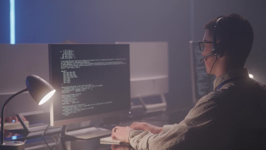 Man stands in front of a computer with codes displayed on the screen wearing a headset in a focused work environment