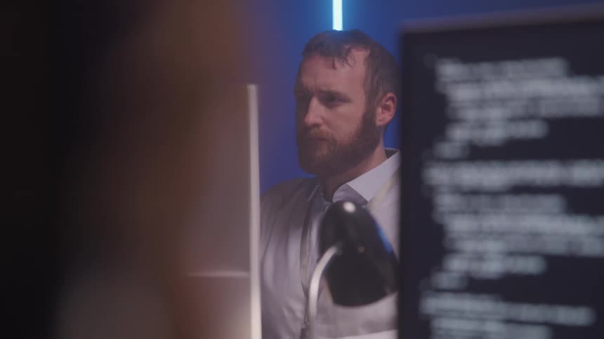 A man works at a desk in a dimly lit room focused on his computer suggesting a call center or tech support setting