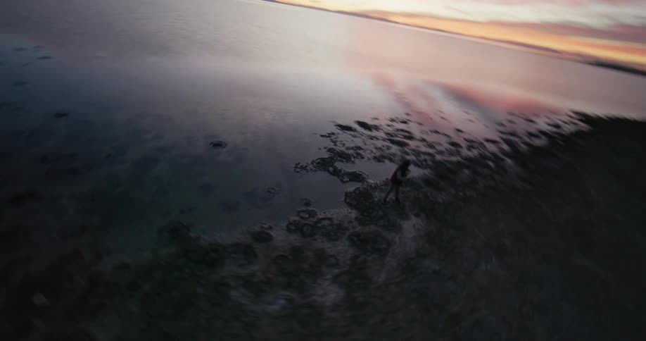 An aerial view of a person dancing on the shore of a large lake with still water and strong sunlight in the sky
