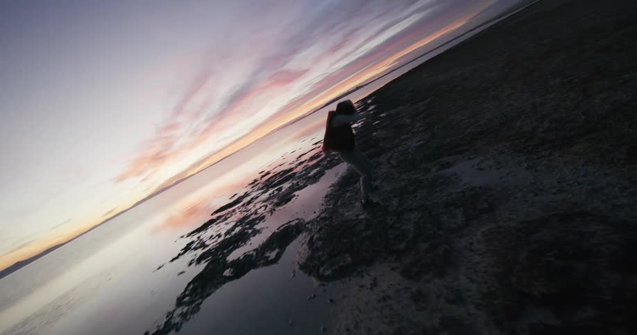 A person dances on a rocky beach nearby and a distant coastline under a sky streaked with clouds