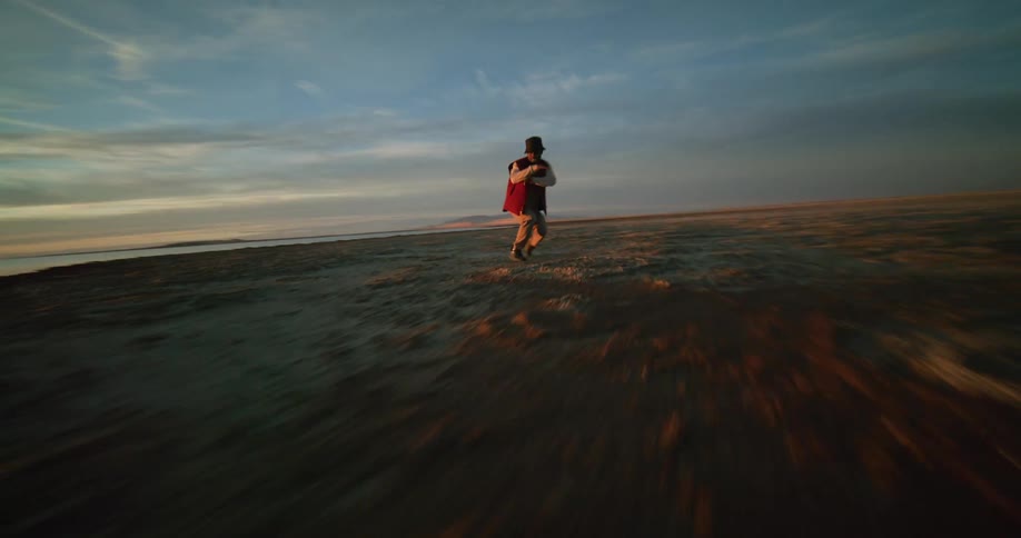 A person dances on a rocky beach nearby and a distant coastline under a sky streaked with clouds