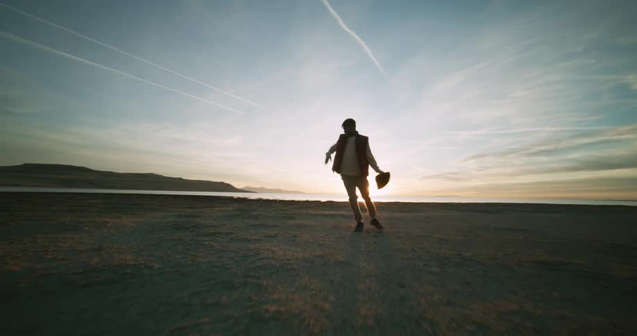 A person dances near a beach with a distant coastline under a sky streaked with clouds at sunset
