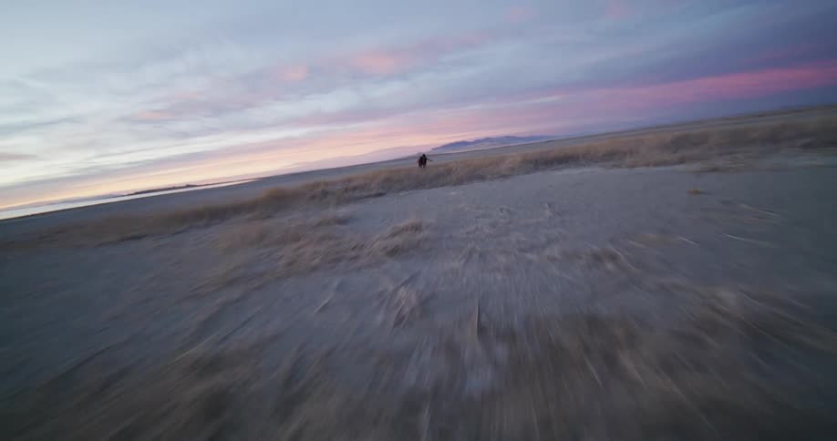 A man wearing a bucket hat and vest dances with arms outstretched in a vast dry grassy field near a calm body of water with distant mountains silhouetted against a sunset sky