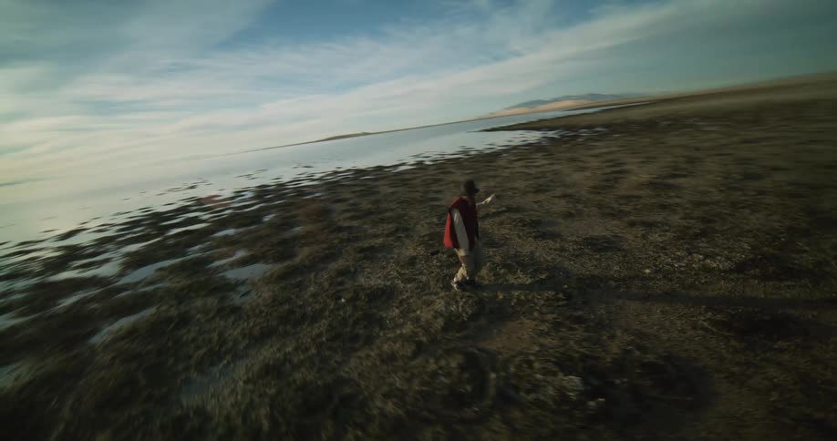 A person dances on a rocky beach nearby and a distant coastline under a sky streaked with clouds