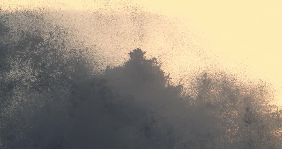 Sea spray bursts against a backdrop of a pale sky at sunset
