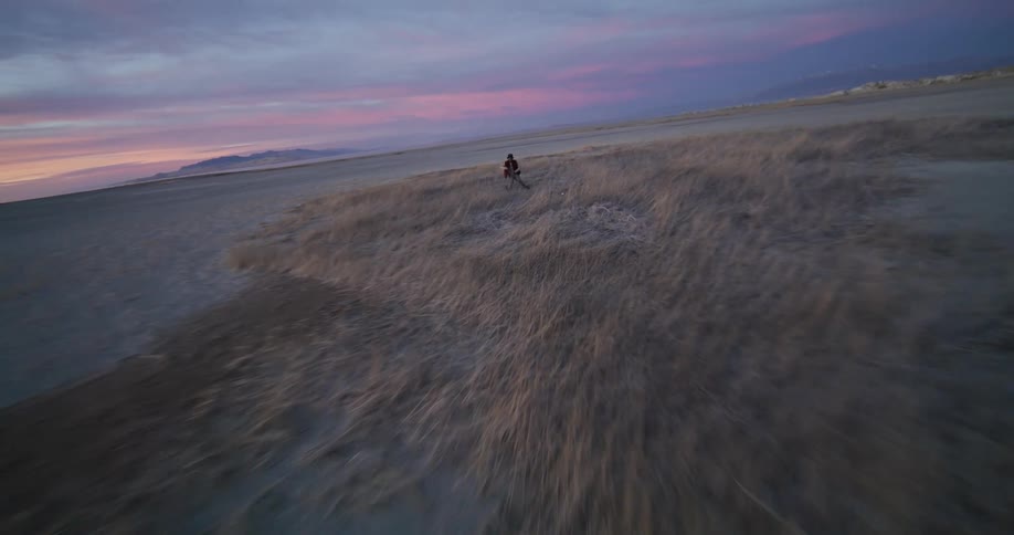 From high above a drone camera descends tracking a person dancing on a grassy hillside as it sweeps past them towards ground level before rising back up into the sky capturing the figure in a red jacket and hat against the coastal sunrise backdrop