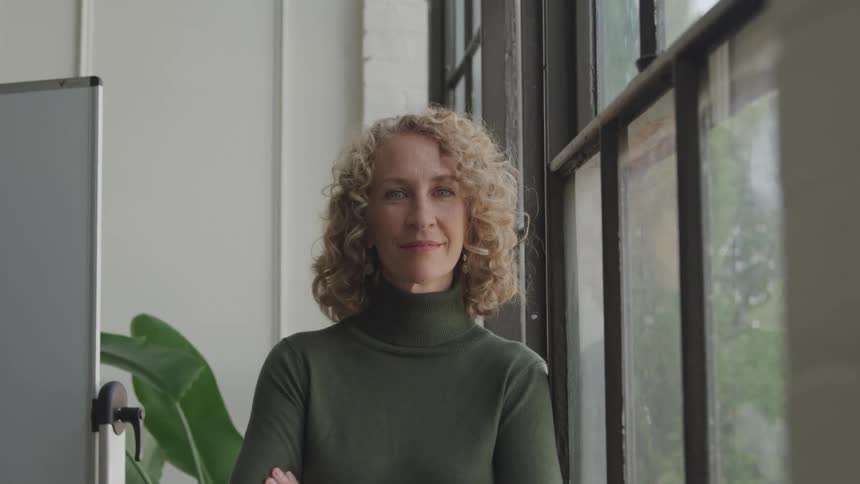 A woman with curly hair wearing a green turtleneck sweater stands beside a window in an office