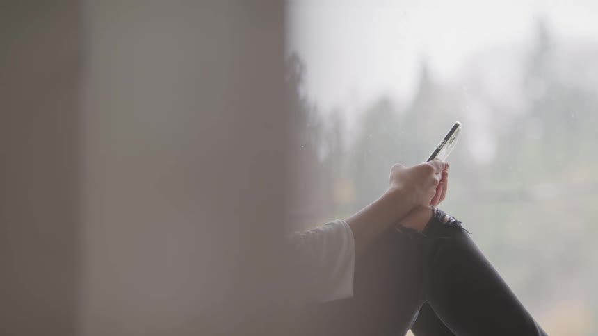 A persons hands are seen holding a phone while texting against a blurred window backdrop
