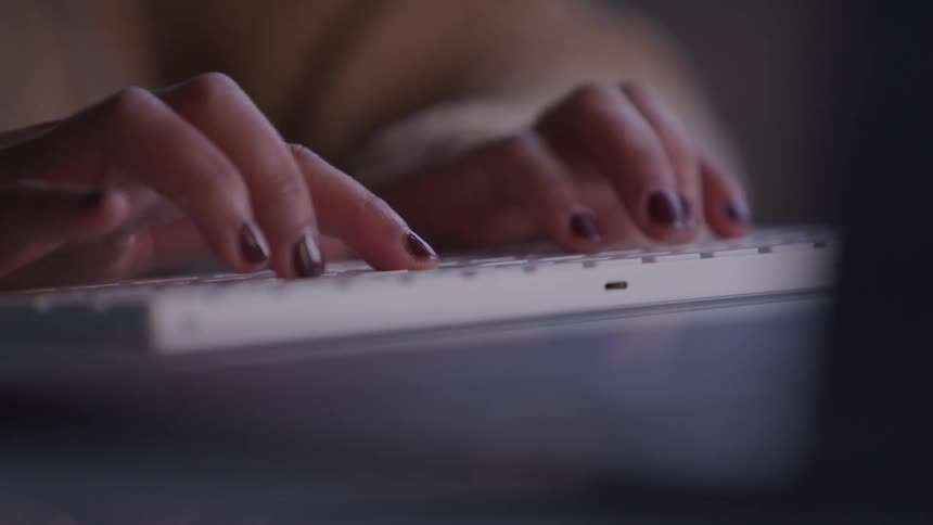Fingers with painted nails type on a white keyboard in a dimly lit setting