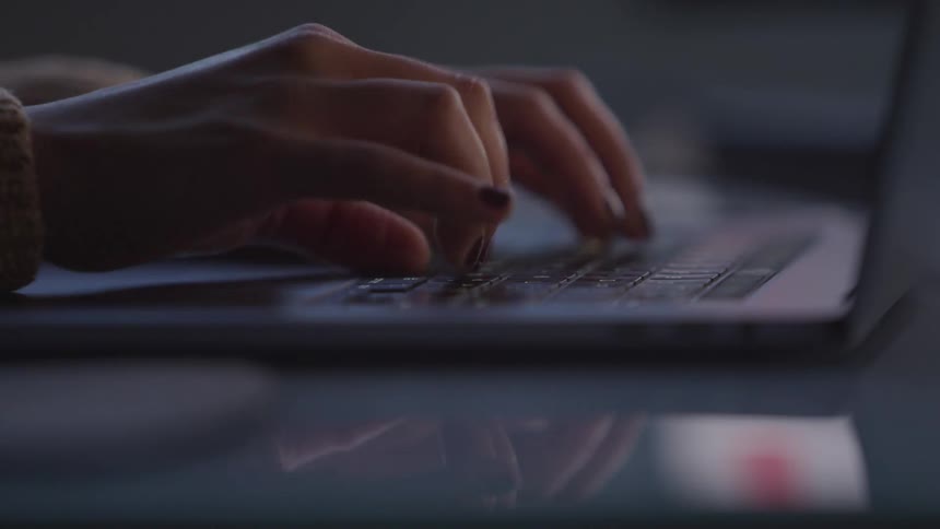 Fingers with dark nail polish type on a white keyboard in a dimly lit setting