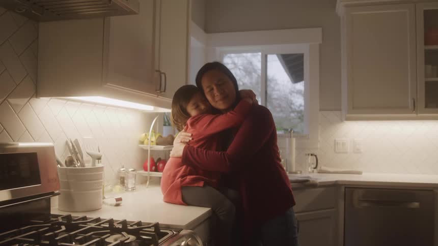 A warmly lit kitchen scene shows a child sitting on the countertop while being embraced by an adult