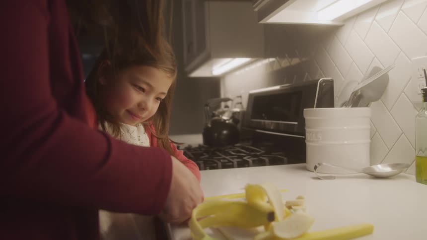 A mother is helping her daughter cut a banana in a home kitchen