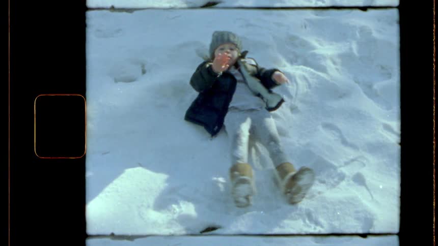 A child lies on the snow dressed in winter clothing including a coat hat and boots