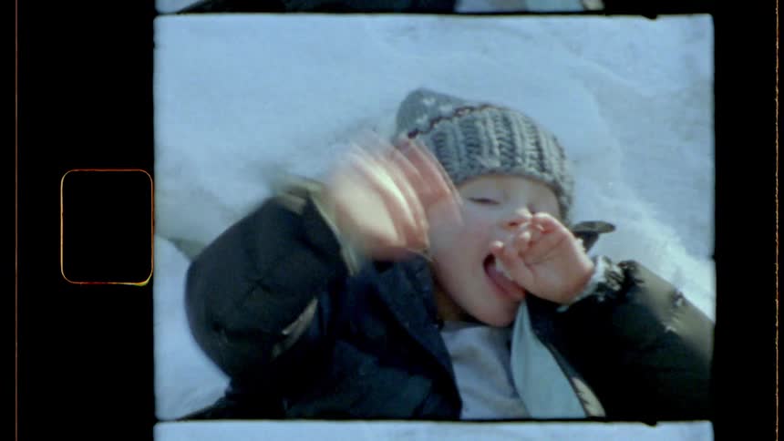 A girl wearing winter clothes lays over a patch of white snow waving at the camera